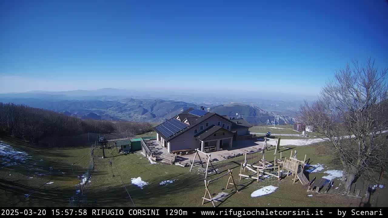 A mountaintop building with solar panels is seen, with a playground and ski lift nearby, under a clear blue sky and patches of snow on the ground.