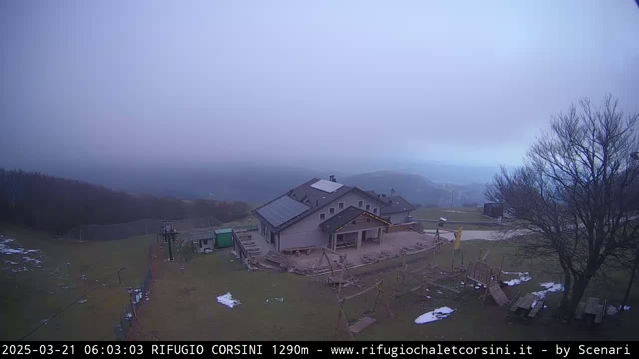 A hazy, overcast day reveals a building with solar panels, playground equipment, and a surrounding landscape of trees and patchy snow.