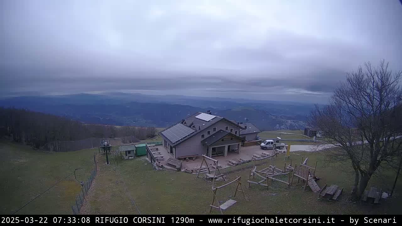 A mountaintop lodge with solar panels sits on a grassy area overlooking a valley under a cloudy sky.