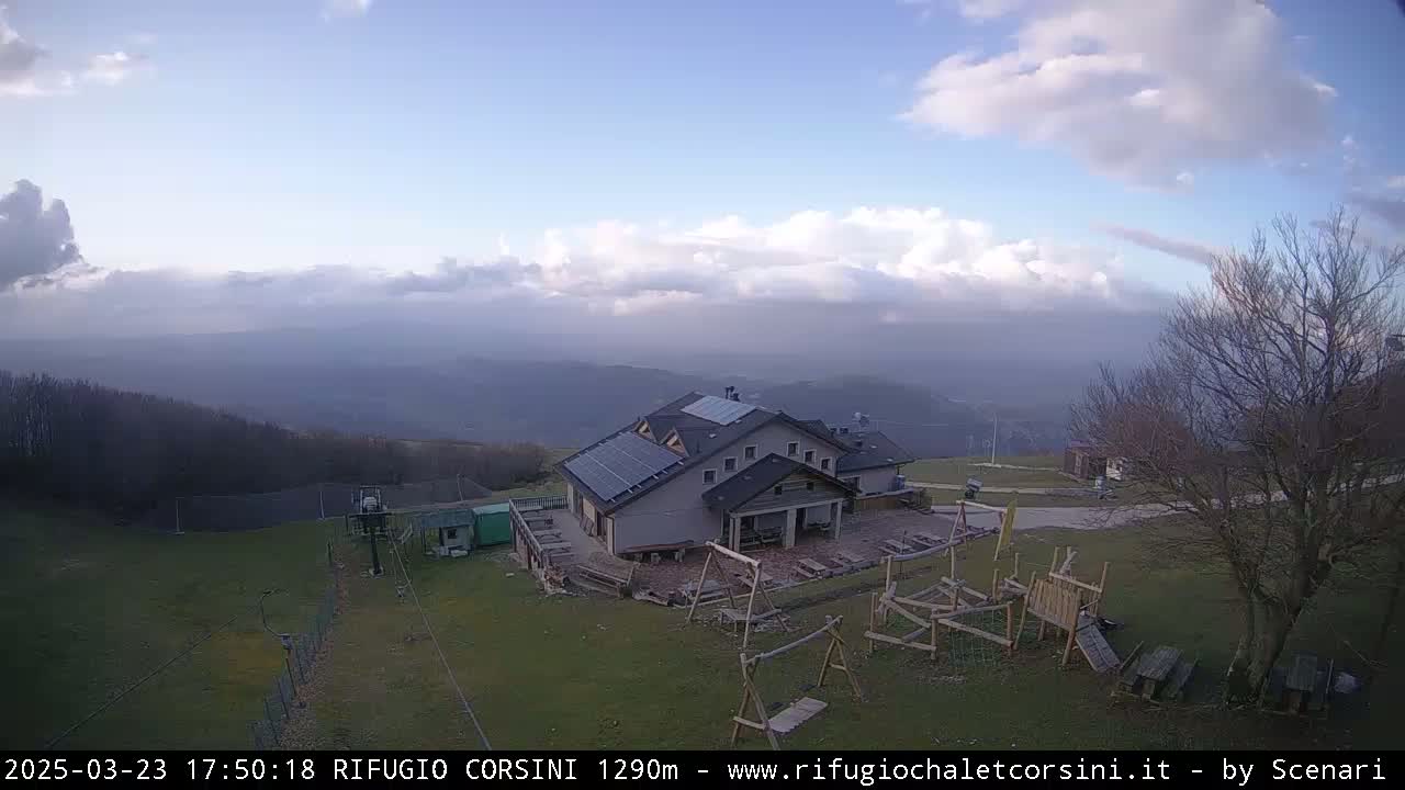 A mountaintop lodge with solar panels sits on grassy terrain overlooking a hazy valley under a partly cloudy sky.
