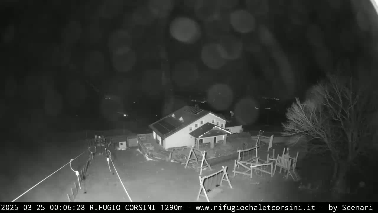 A nighttime, rainy view of a building with a playground and some bare trees in the foreground.