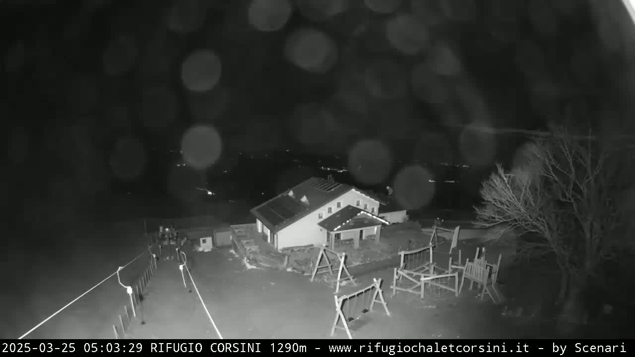 A nighttime, rain-blurred view of a building with a playground next to it, and a few people walking on a road in the foreground.