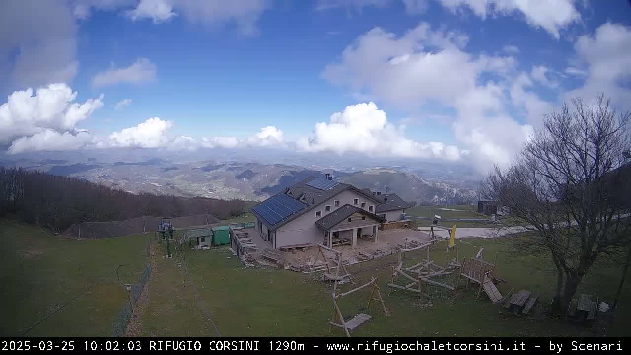 A mountaintop lodge with solar panels sits on a grassy area with playground equipment, overlooking a valley under a partly cloudy blue sky.