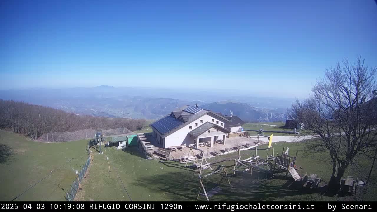 A mountaintop building with solar panels is seen on a sunny day, overlooking a vast valley and distant mountains.
