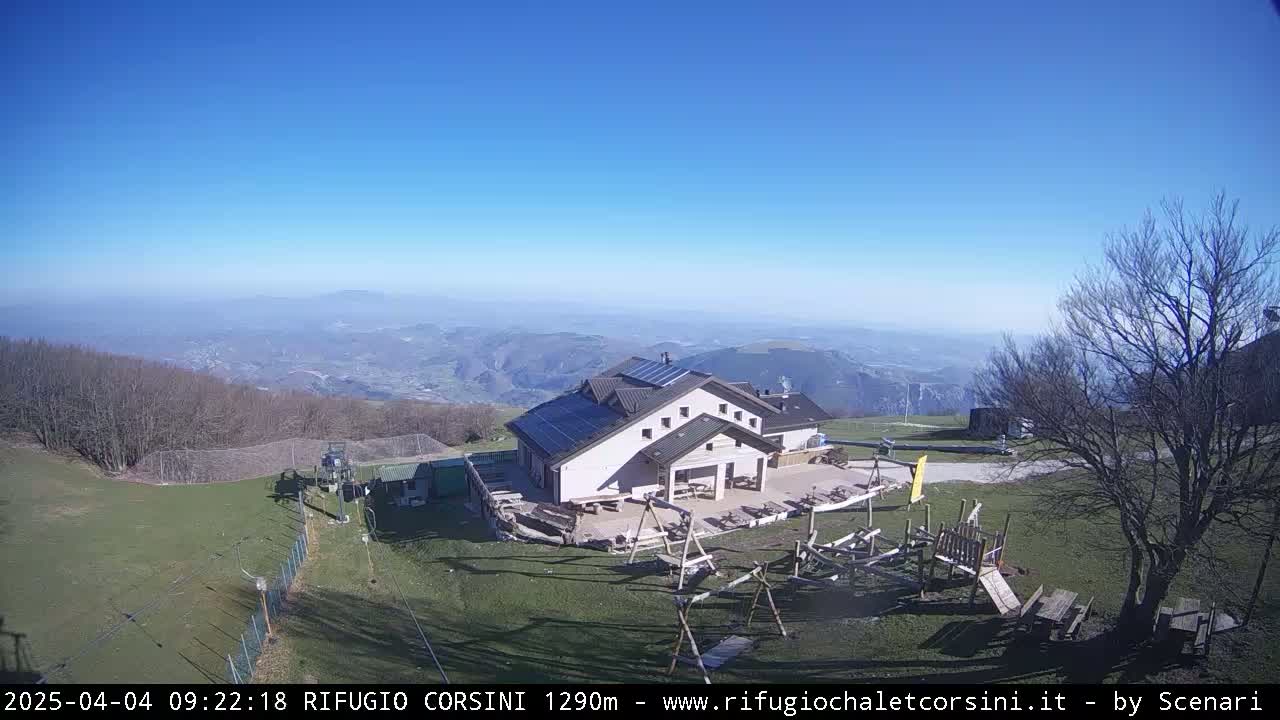 A mountaintop building with solar panels on its roof, a playground, and surrounding grassy areas, overlooks a hazy valley under a clear blue sky.