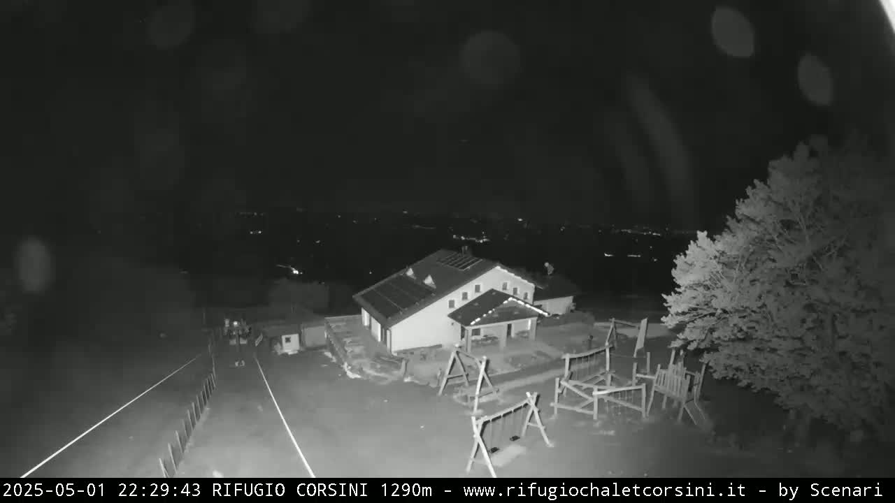 A nighttime, high-angle view shows a building with solar panels on its roof, a playground, and a road leading to a dimly lit town in the distance.