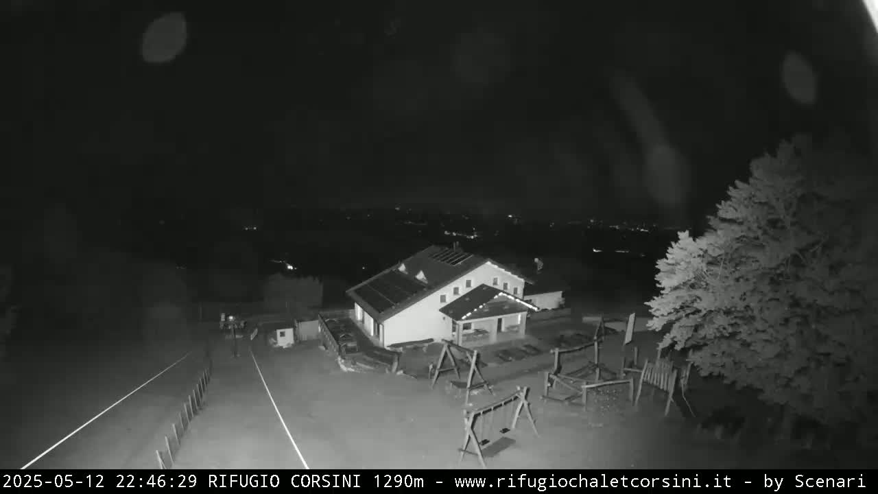 A nighttime, low-light image shows a building with solar panels on its roof, a playground, and some trees, all situated on a hill overlooking a distant, dimly lit town.