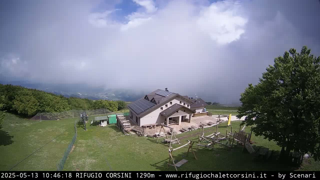 A mountain lodge with solar panels on the roof sits on a grassy area with a playground, under a partly cloudy sky with a hint of fog or mist in the distance.