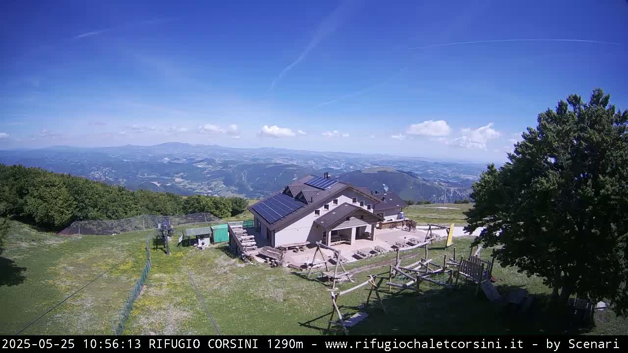 A mountaintop lodge with solar panels sits on a grassy area overlooking a vast, sunny valley.