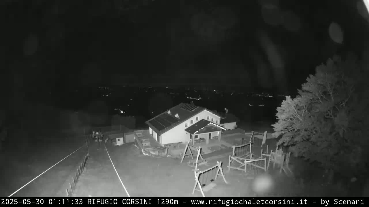 A nighttime, black and white image shows a building with solar panels, a playground, and a roadway leading to a distant, dimly lit town under a clear sky.