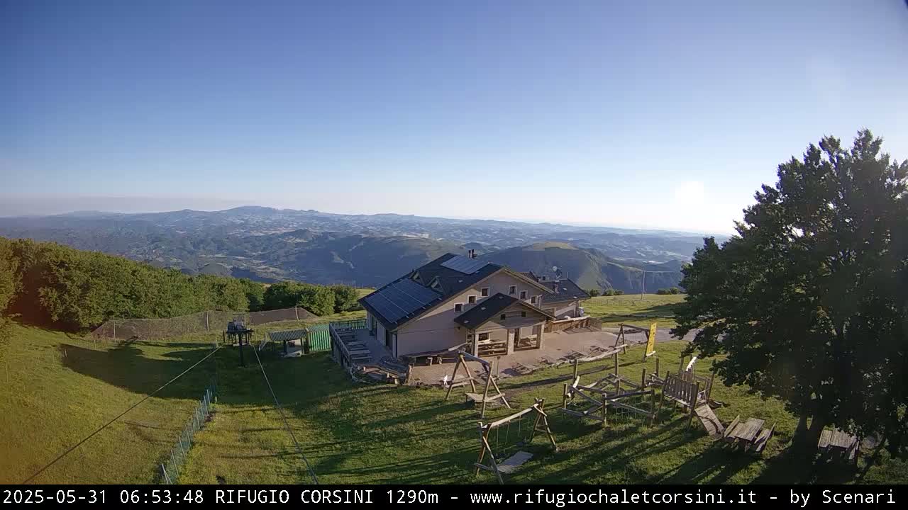 A mountaintop building with solar panels and a playground overlooks a vast, hazy valley under a clear blue sky.