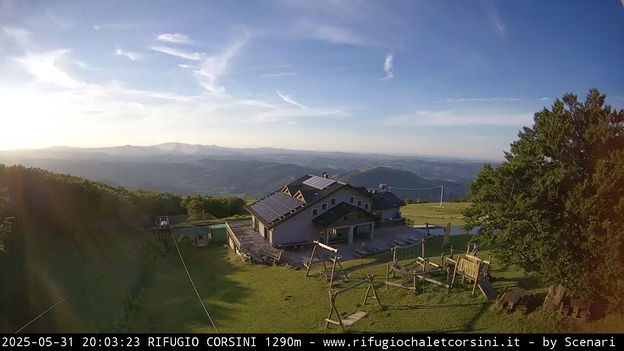 A mountain lodge with solar panels sits on a grassy area with playground equipment, overlooking a vast, hazy valley under a mostly clear sky.