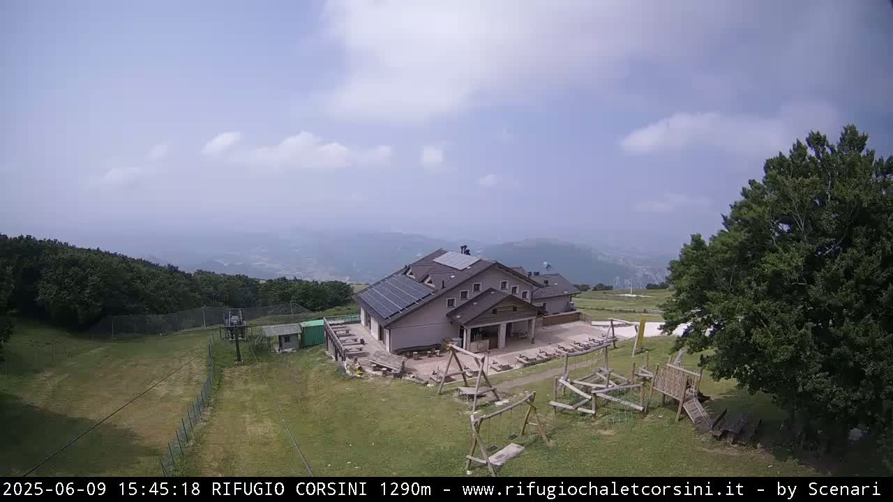 A mountain lodge with solar panels sits on a grassy area with playground equipment, overlooked by a partly cloudy sky and distant hazy mountains.