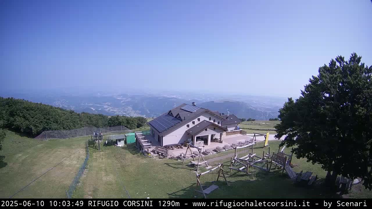 A mountaintop building with solar panels is shown, overlooking a valley on a clear, sunny day; playground equipment and outdoor seating are nearby.