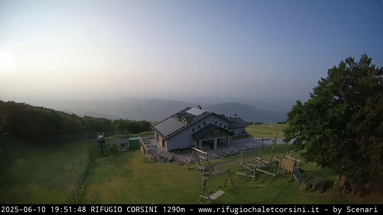 A mountain lodge with solar panels on its roof sits on a grassy area with a playground, overlooking hazy hills and a forest under a clear, pale sky.