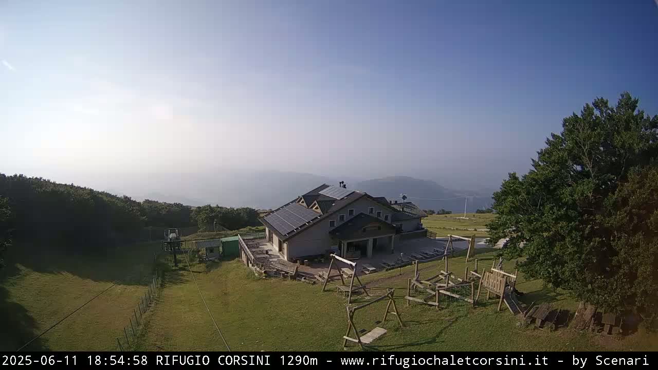 A mountain lodge with solar panels on the roof sits on a grassy area with a playground, overlooking hazy hills and a valley under a clear, sunny sky.