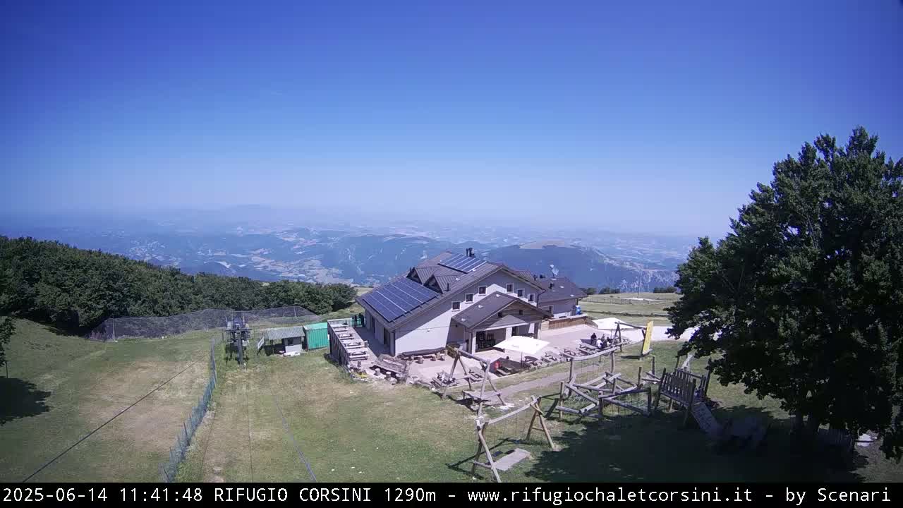 A mountaintop lodge with solar panels sits on a grassy area with a playground, overlooking a vast, hazy valley under a clear blue sky.