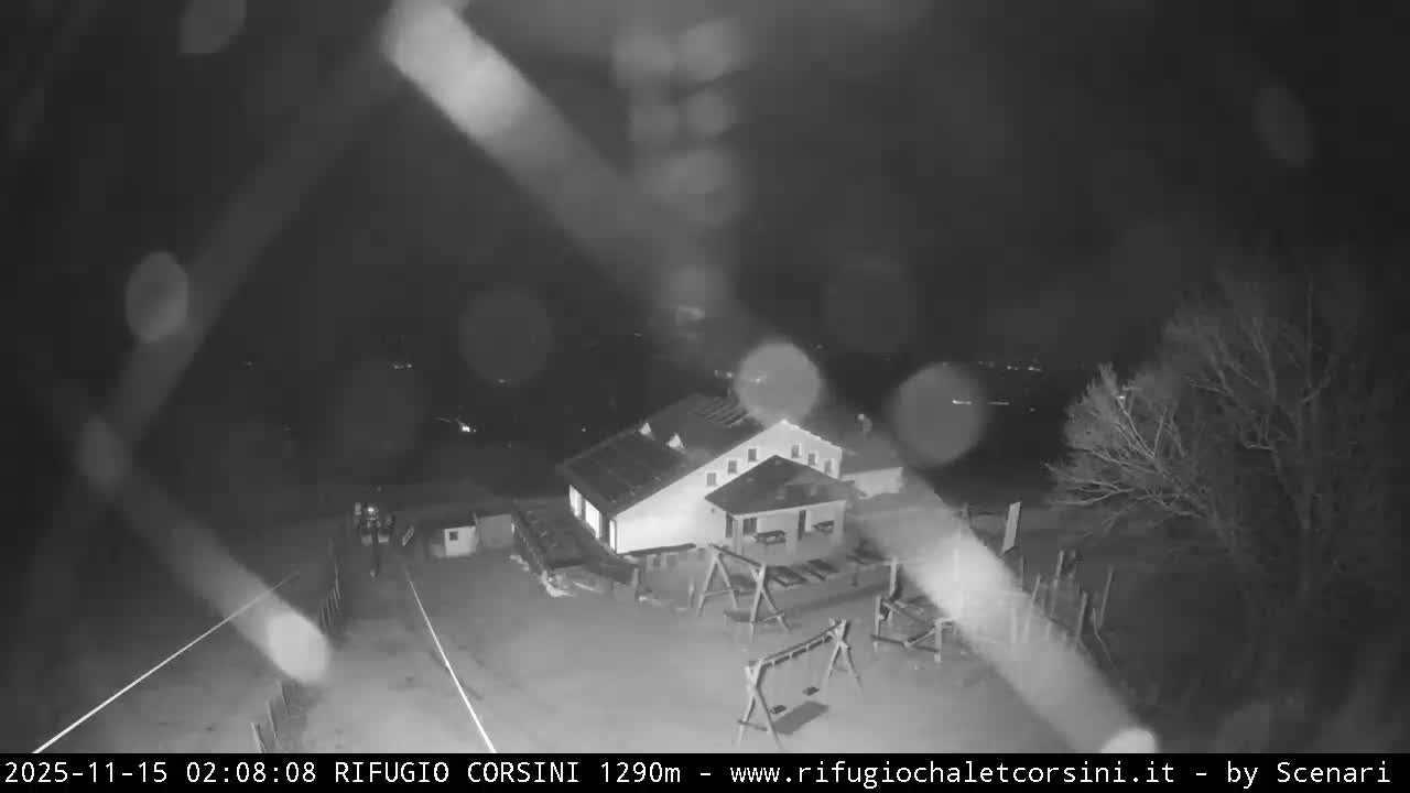 A black and white night-vision image captures a mountain lodge and a playground with bare trees outdoors, viewed through a camera lens blurred by light precipitation, with distant lights faintly visible in the background.