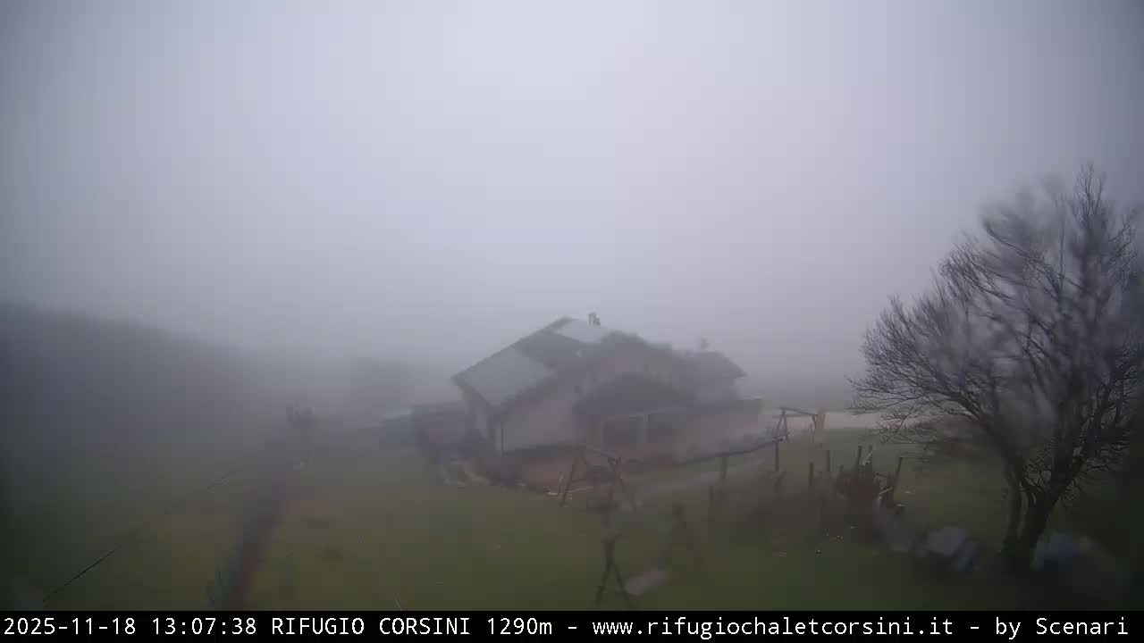 A black and white night-vision image captures a mountain lodge and a playground with bare trees outdoors, viewed through a camera lens blurred by light precipitation, with distant lights faintly visible in the background.