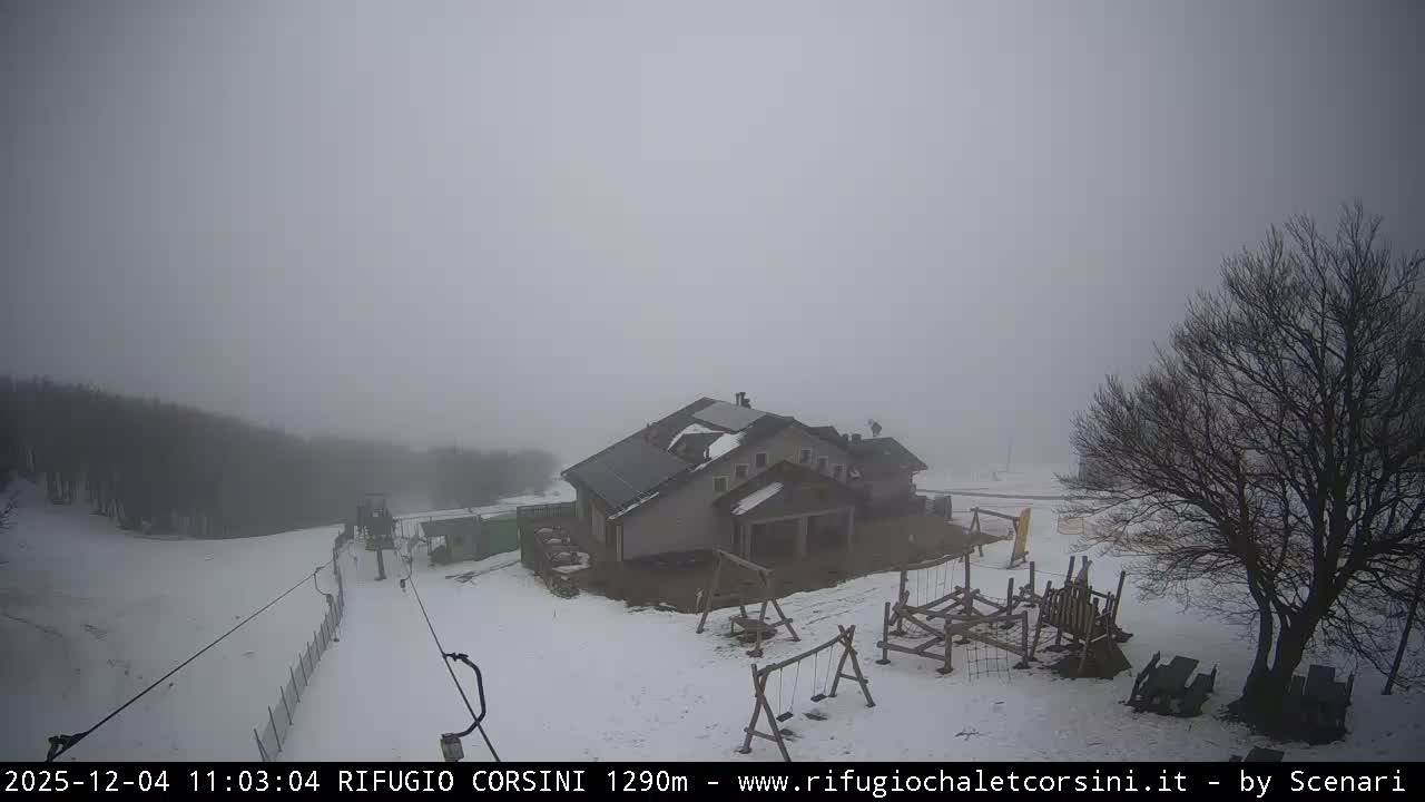 A snow-covered ski resort area, featuring a large lodge with solar panels, a ski lift, and wooden playground equipment, is enveloped in thick fog under overcast skies.