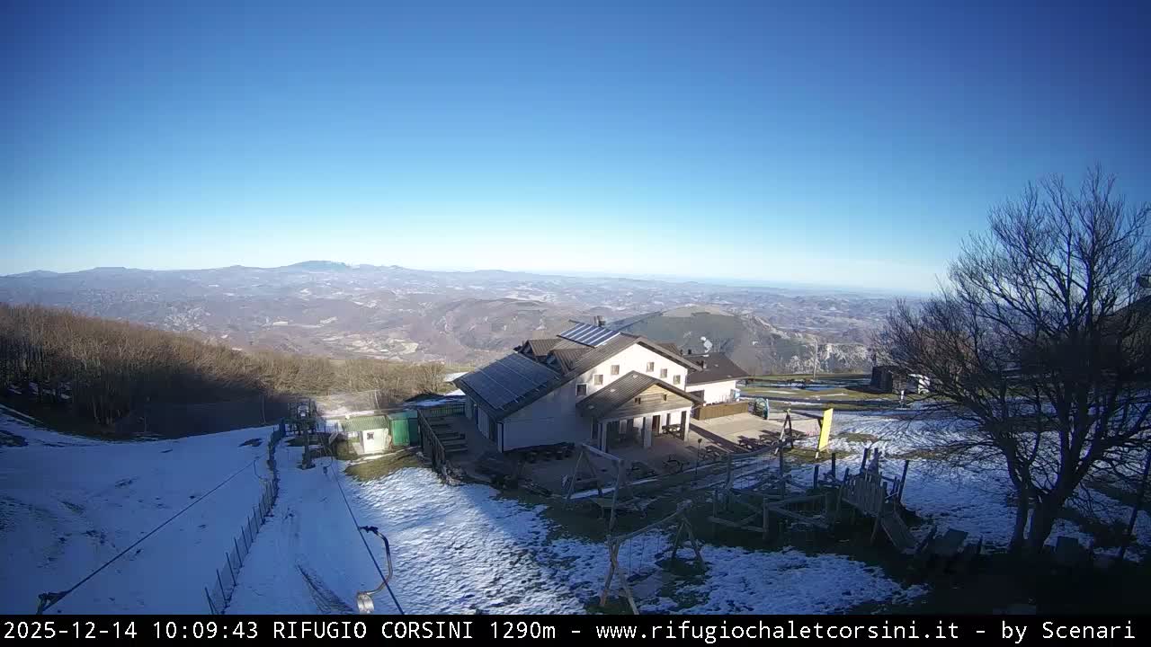 A snow-covered ski resort area, featuring a large lodge with solar panels, a ski lift, and wooden playground equipment, is enveloped in thick fog under overcast skies.