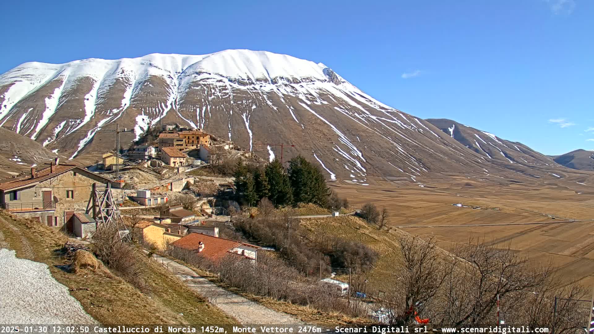 A small village nestled in a valley below a snow-capped mountain on a sunny day.