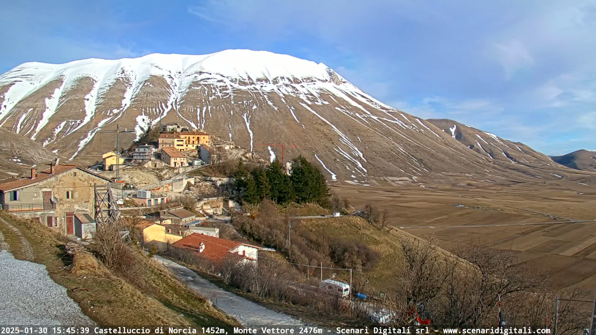 A small village nestled in a valley, with a snow-capped mountain in the background under a mostly sunny sky.