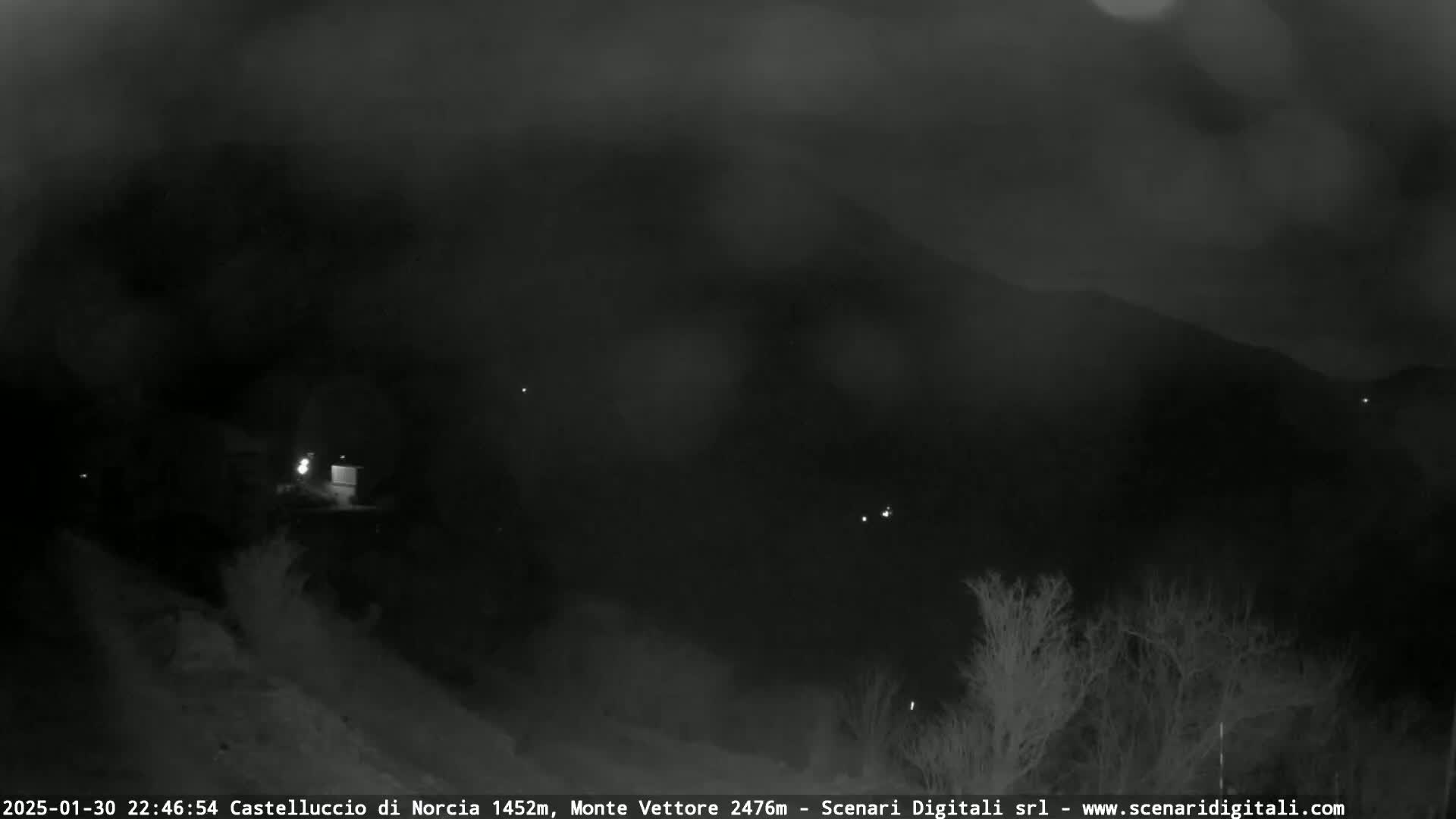 A nighttime, low-light view of a dark landscape with sparsely scattered lights and silhouetted trees under a cloudy sky.