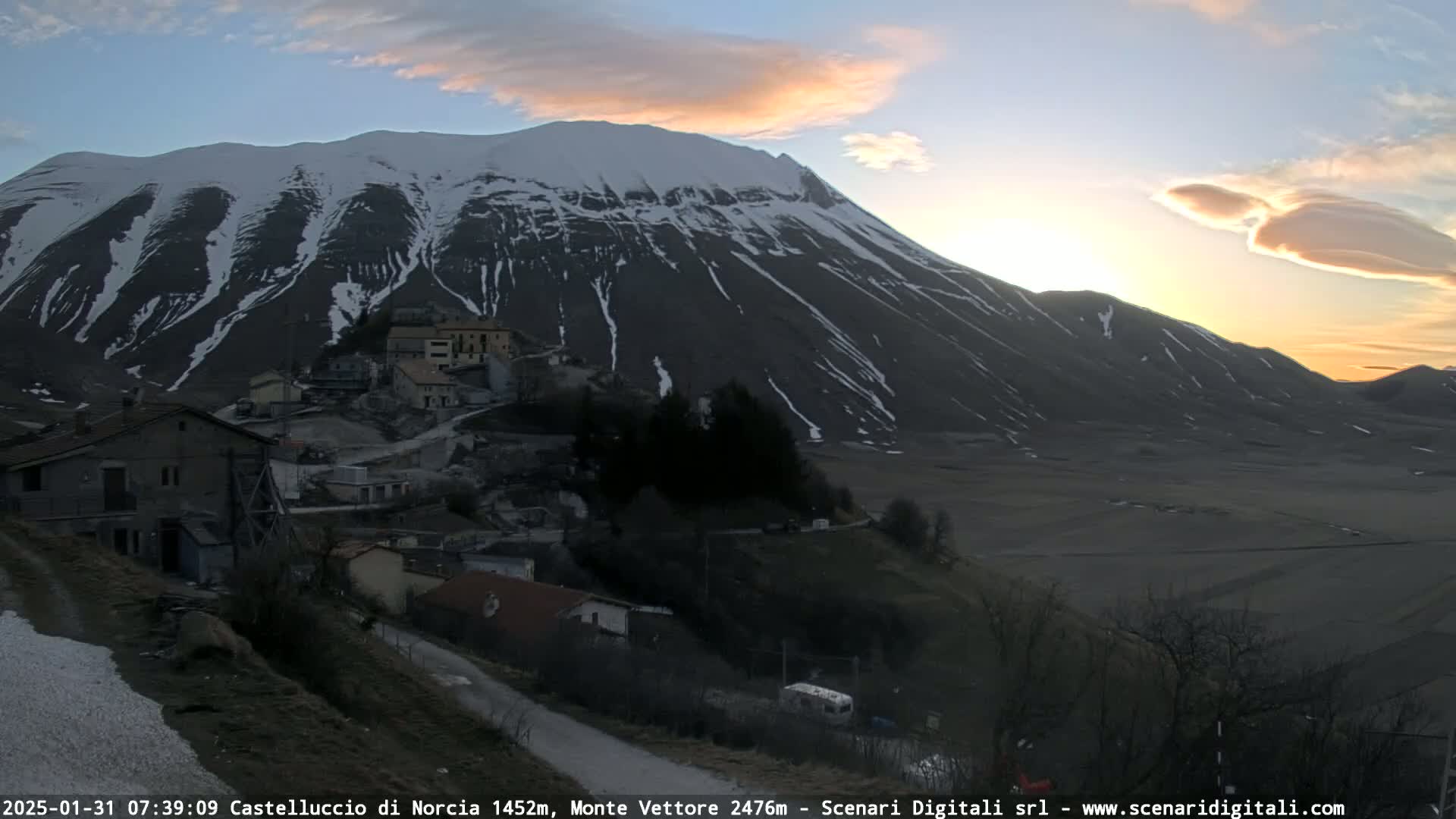 A snow-dusted mountain overlooks a small village at sunset under a partly cloudy sky.