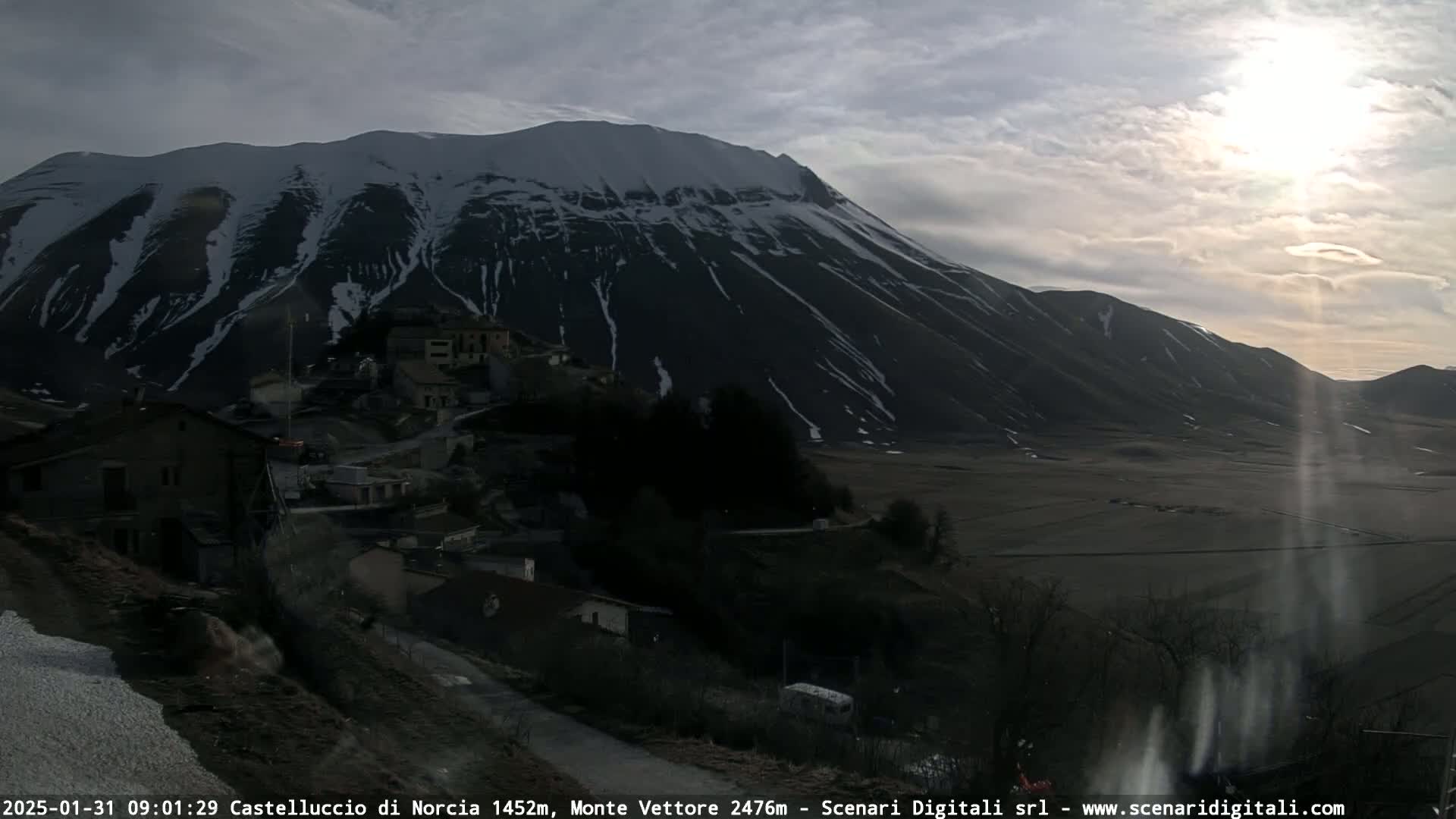 A snow-dusted mountain looms over a small village nestled in a valley under a partly cloudy sky, with the sun shining brightly.