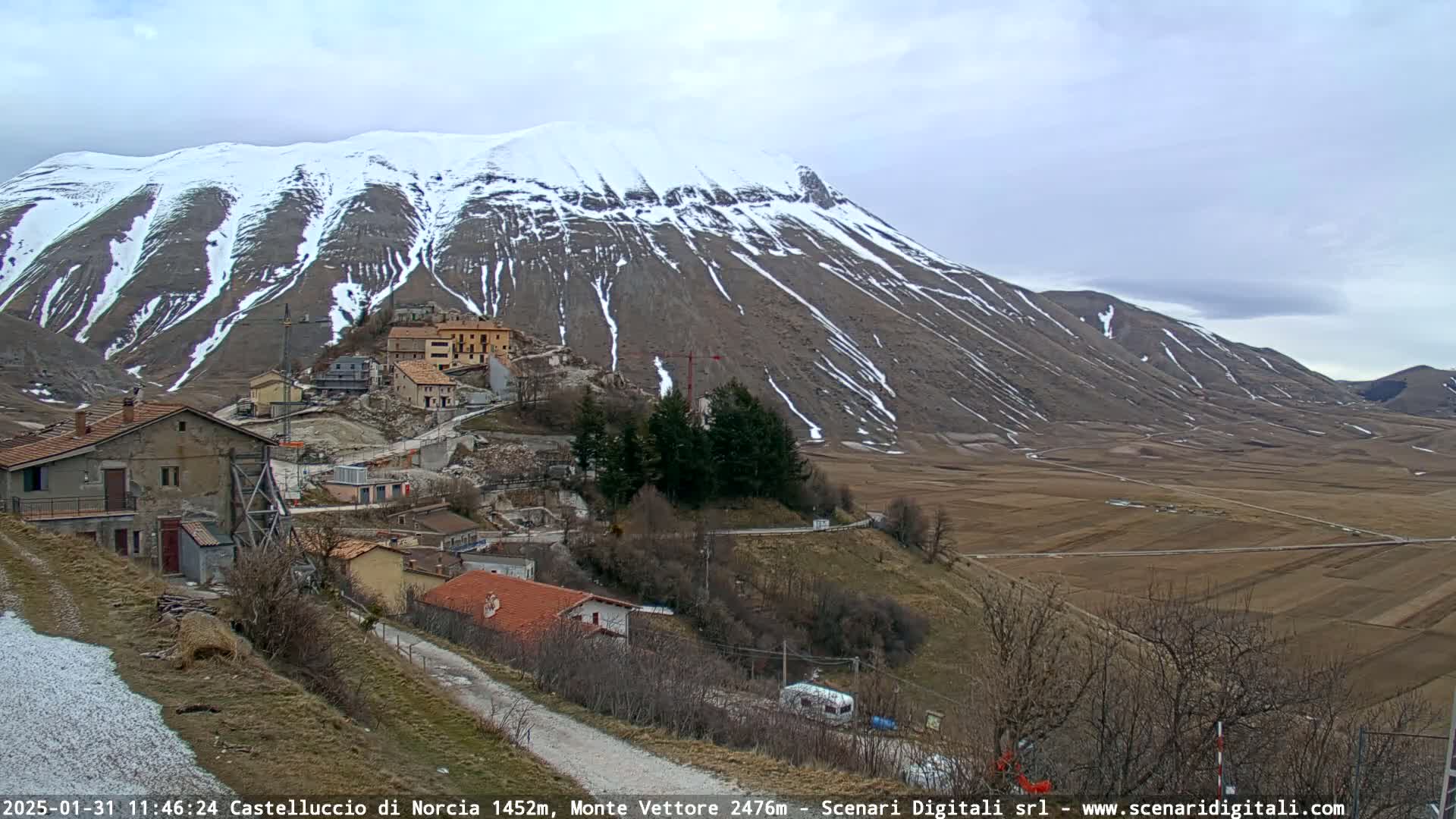 A snow-dusted mountain overlooks a small village nestled in a valley of brown fields under a cloudy sky.