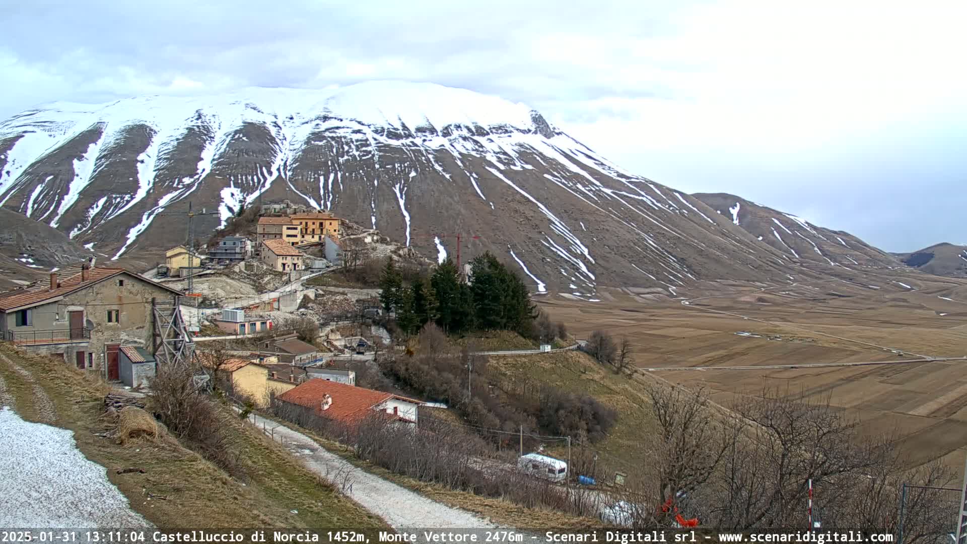 A snow-dusted mountain overlooks a small village nestled in a valley of brown fields under a mostly cloudy sky.