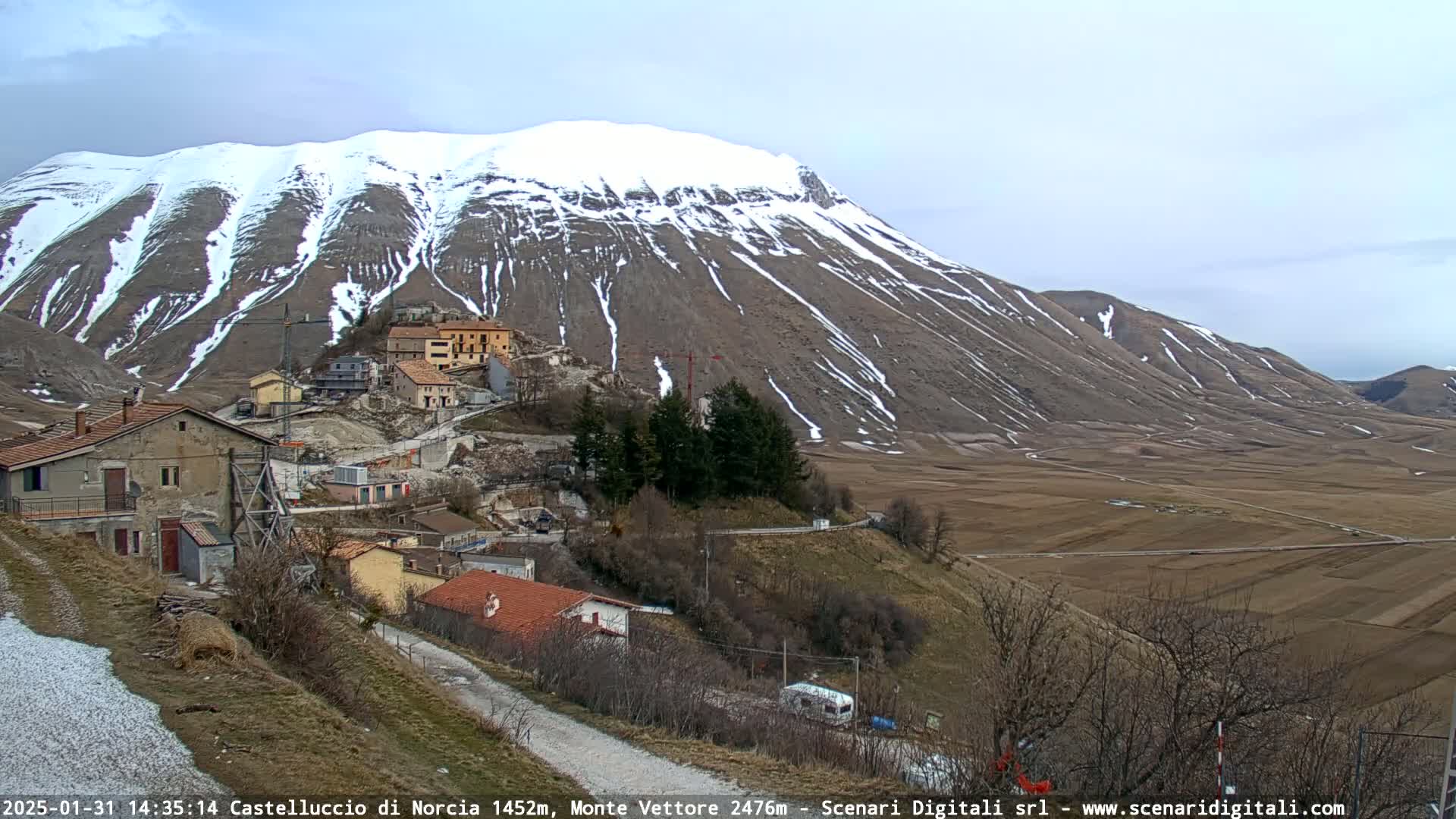 A snow-dusted mountain overlooks a small village nestled in a valley of brown fields under a cloudy sky.