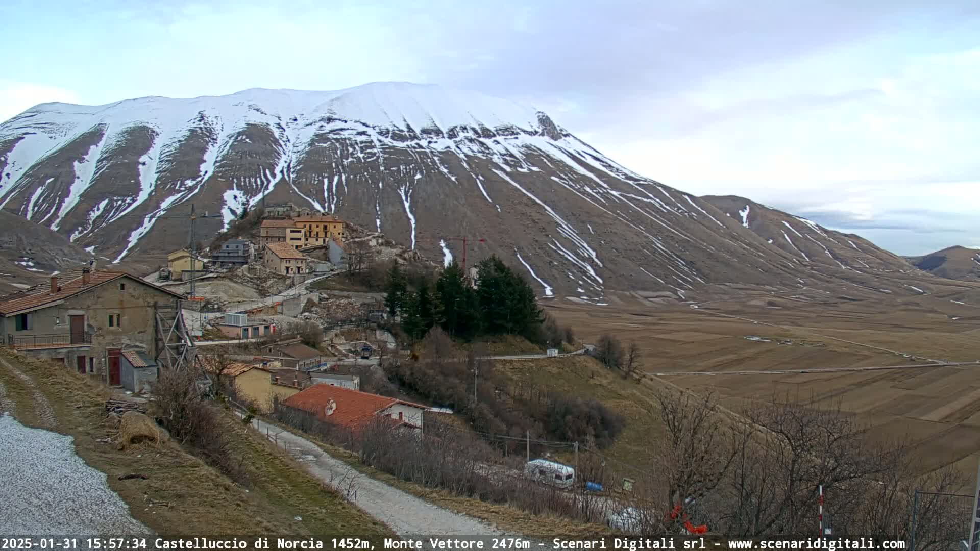 A snow-dusted mountain overlooks a small village nestled in a valley of brown fields under a cloudy sky.