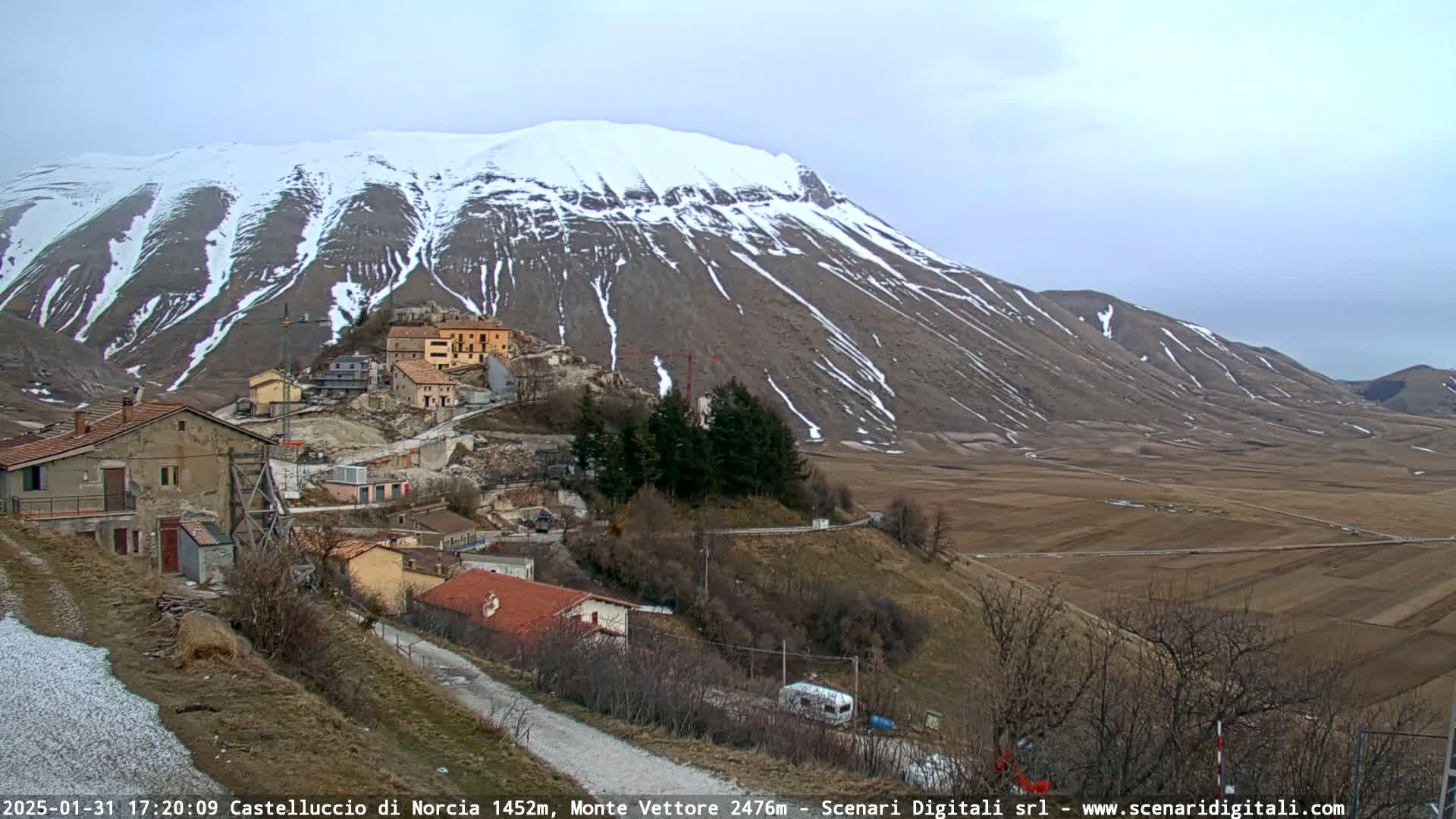 A small village nestled in a valley below a snow-dusted mountain range on an overcast day.