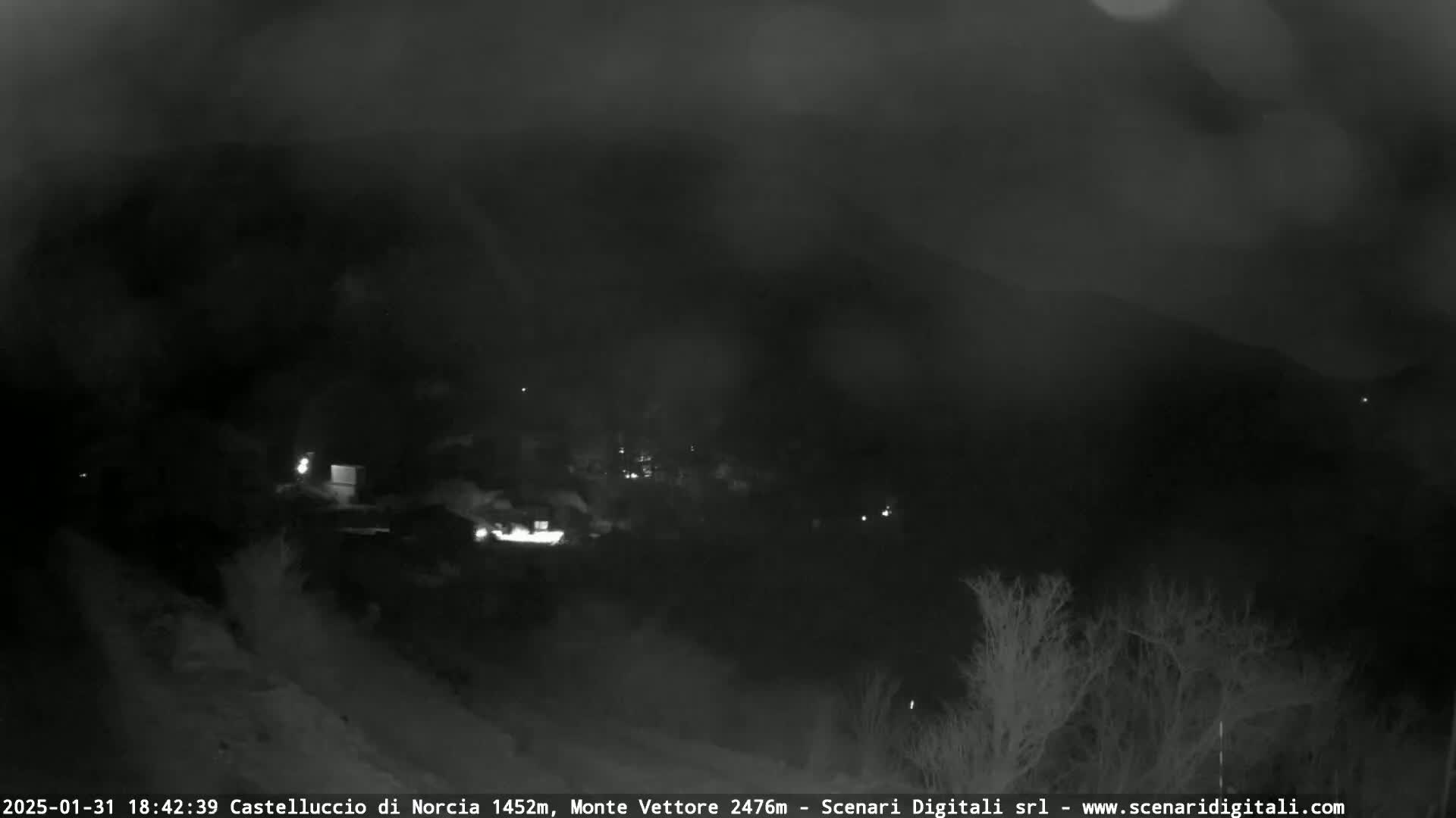 A nighttime, low-visibility view of a dimly lit town nestled in a valley, partially obscured by fog or mist.