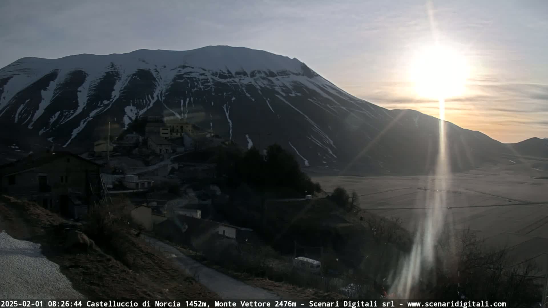 A sunlit, snow-capped mountain overlooks a small village nestled in a valley at sunrise.