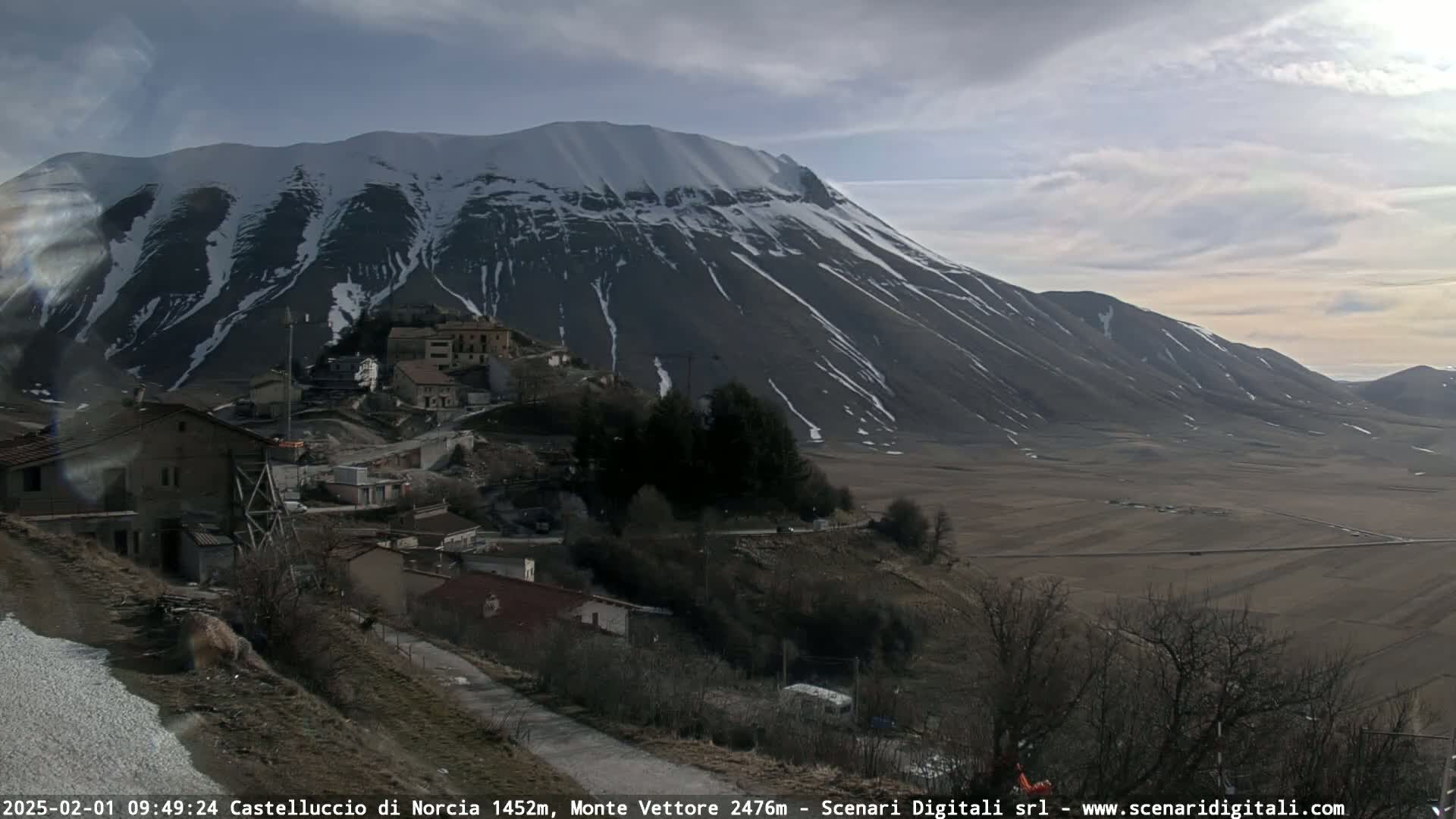 A snow-capped mountain overlooks a small village nestled in a valley under a partly cloudy sky.