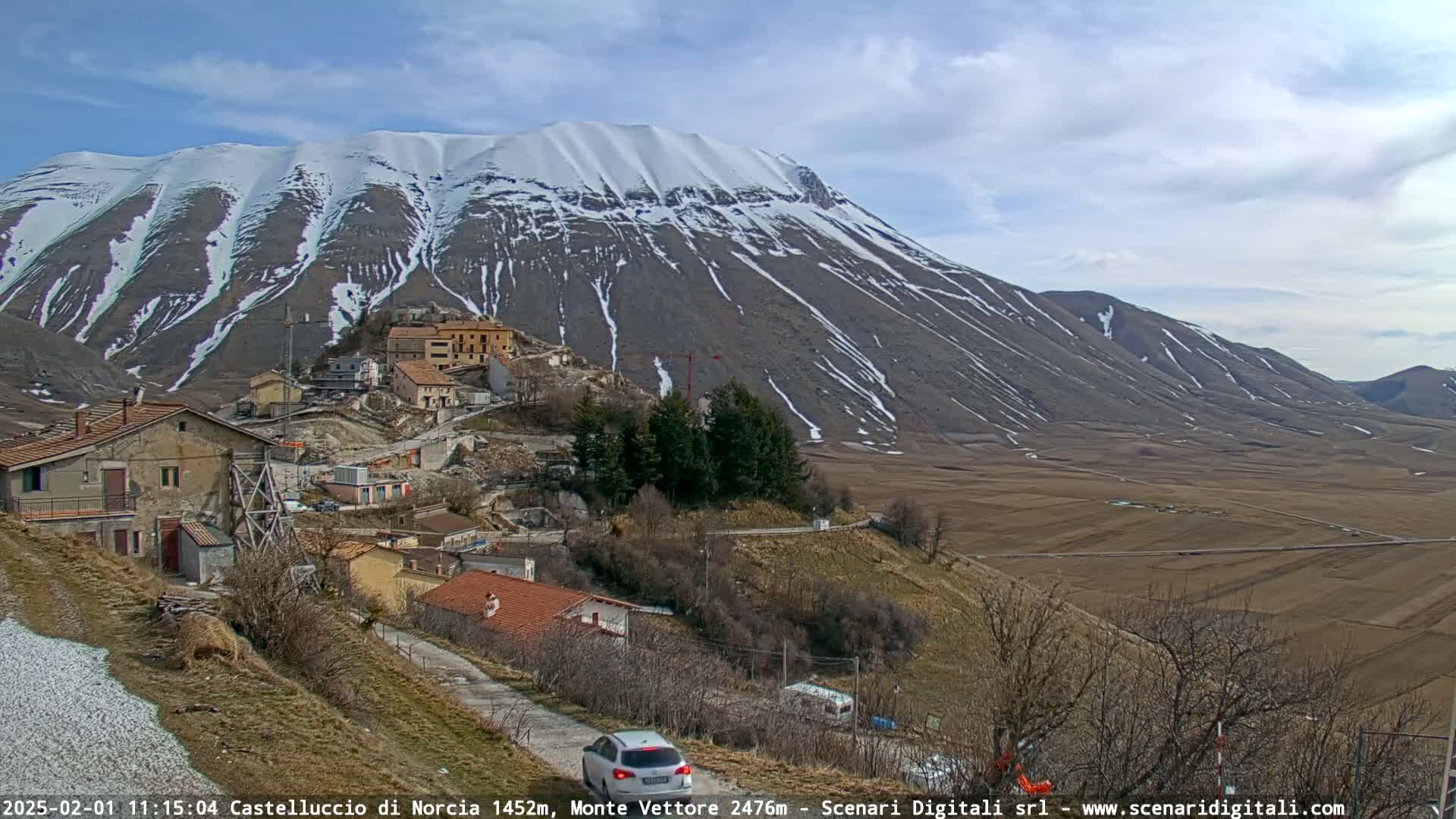 A small village nestled in a valley, with a snow-capped mountain in the background under a partly cloudy sky.