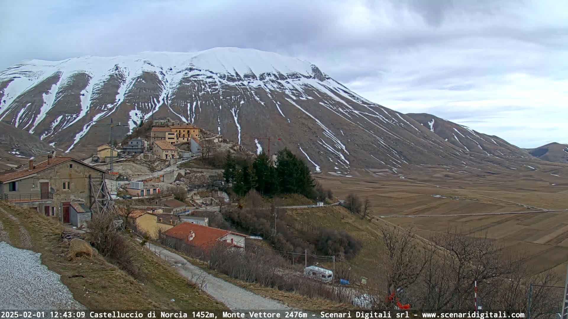 A snow-dusted mountain overlooks a small village nestled in a valley under a cloudy sky.