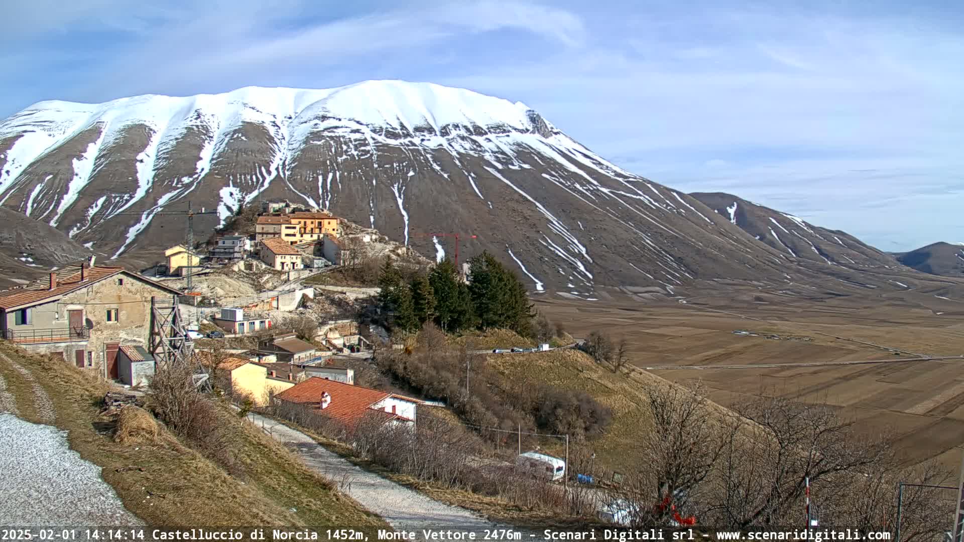 A snow-dusted mountain overlooks a small village nestled in a valley of brown fields under a clear blue sky.