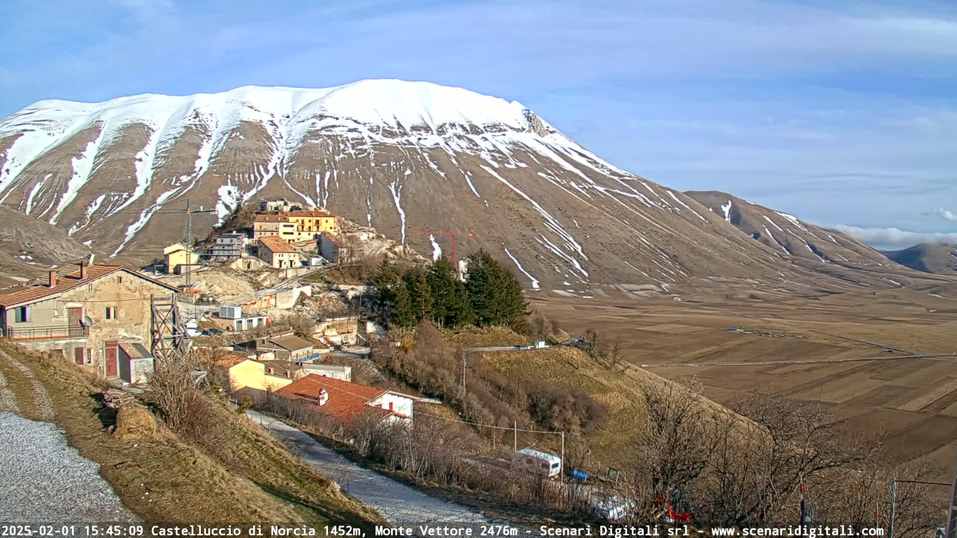 A snow-capped mountain overlooks a small village nestled in a valley of brown fields under a clear blue sky.