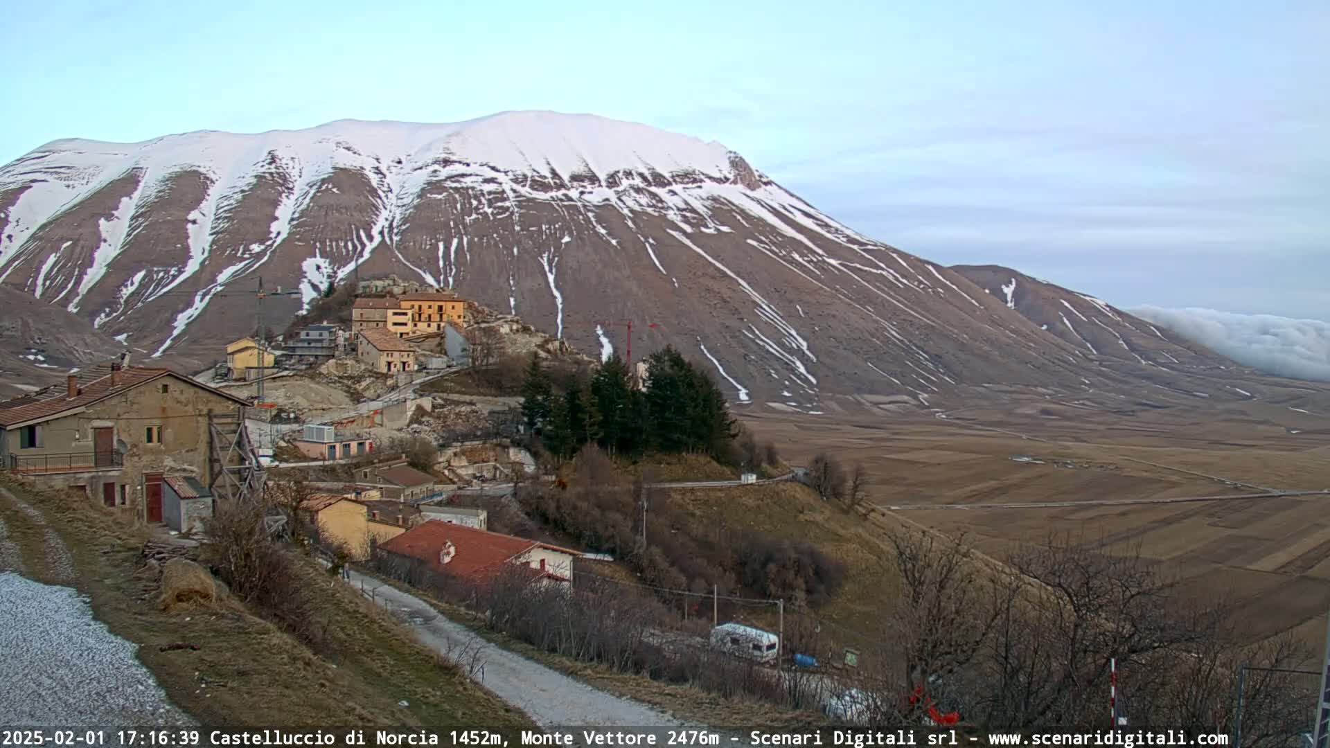 A snow-dusted mountain overlooks a small village nestled in a valley, under a mostly clear sky with some low clouds in the distance.