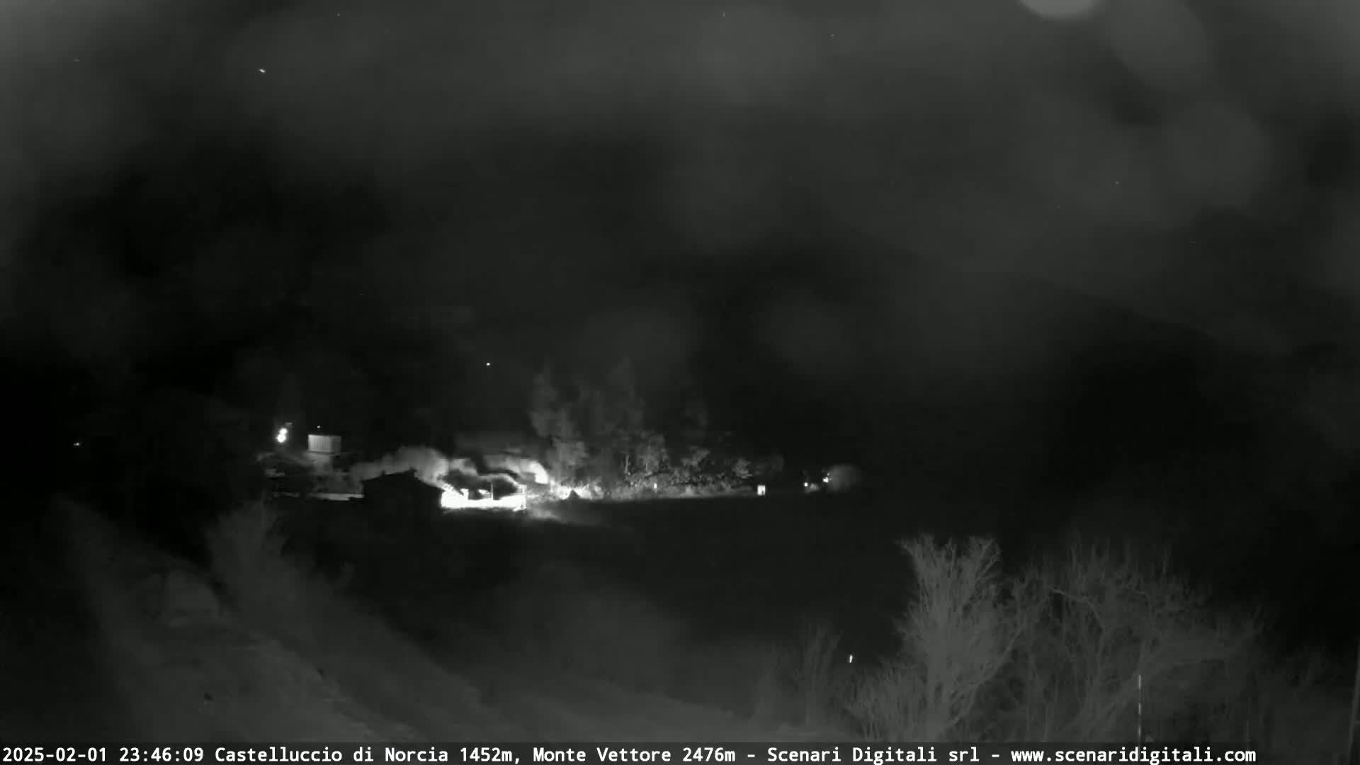 A nighttime, low-light image shows a fire burning in a wooded area.