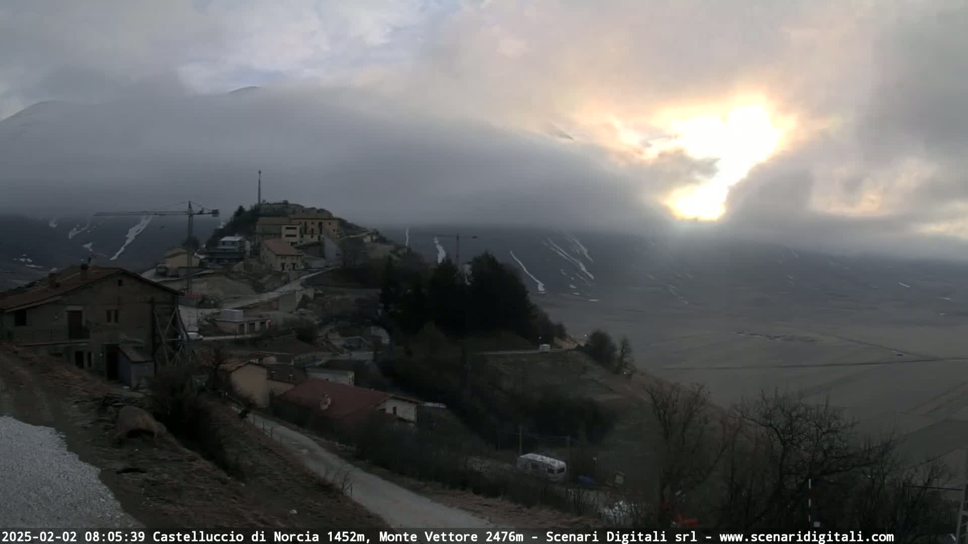 A sunlit, foggy morning reveals a small mountain village with a construction crane, nestled below a snow-dusted mountain range.