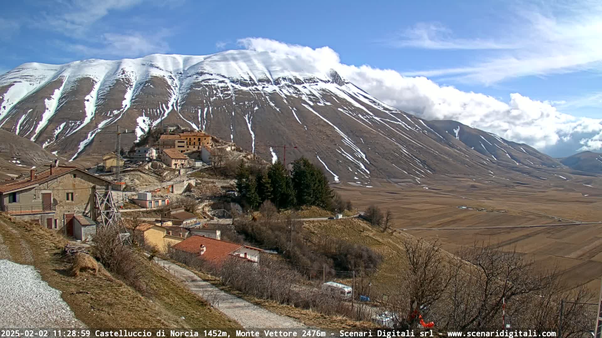 A partially snow-covered mountain looms over a small village nestled in a valley under partly cloudy skies.