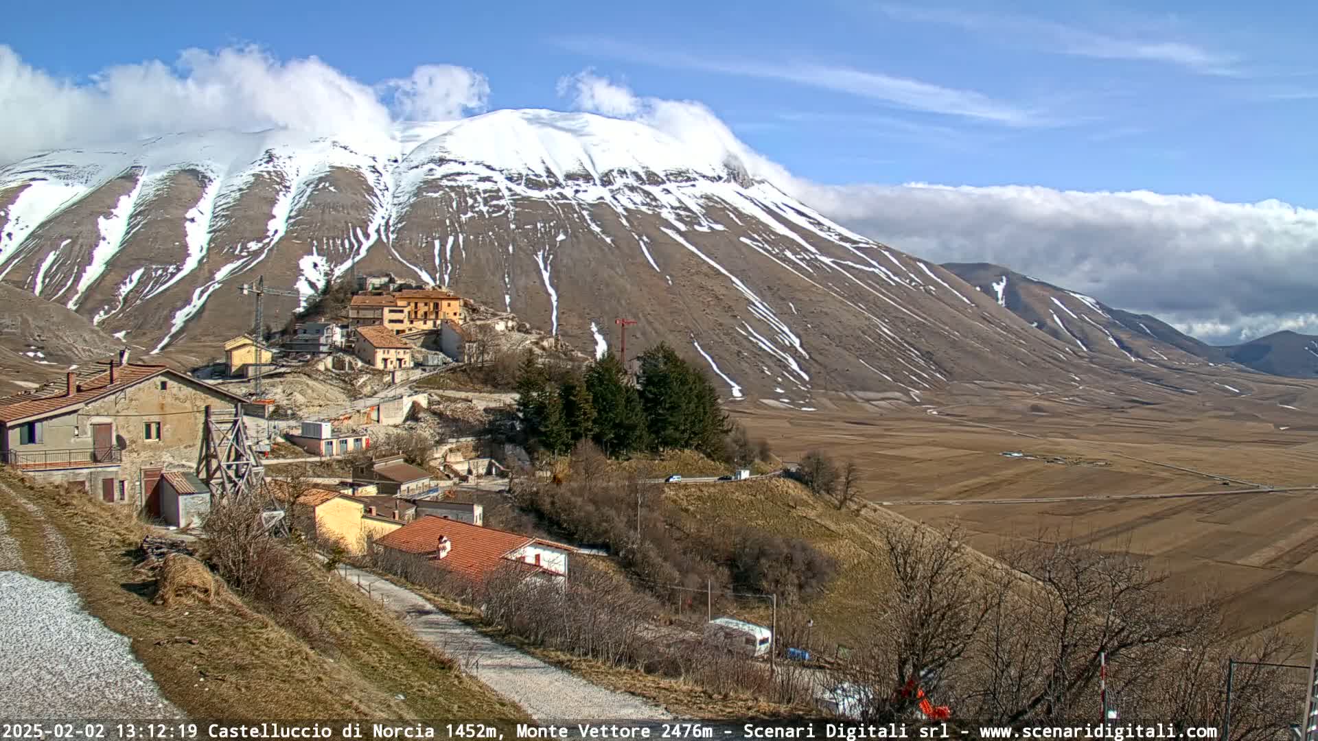 A small village nestled in a valley, with a snow-capped mountain in the background under a partly cloudy sky.