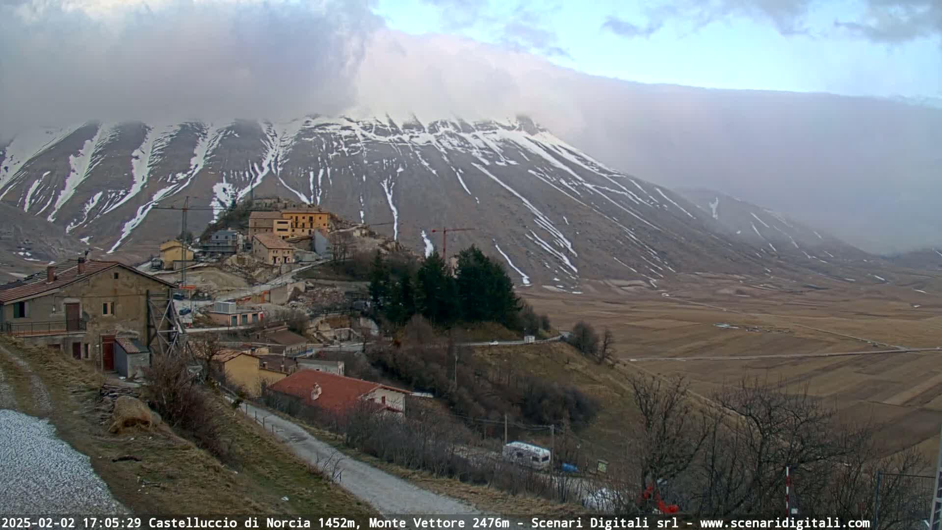 A small village nestled in a valley below a large, snow-dusted mountain range under an overcast sky.