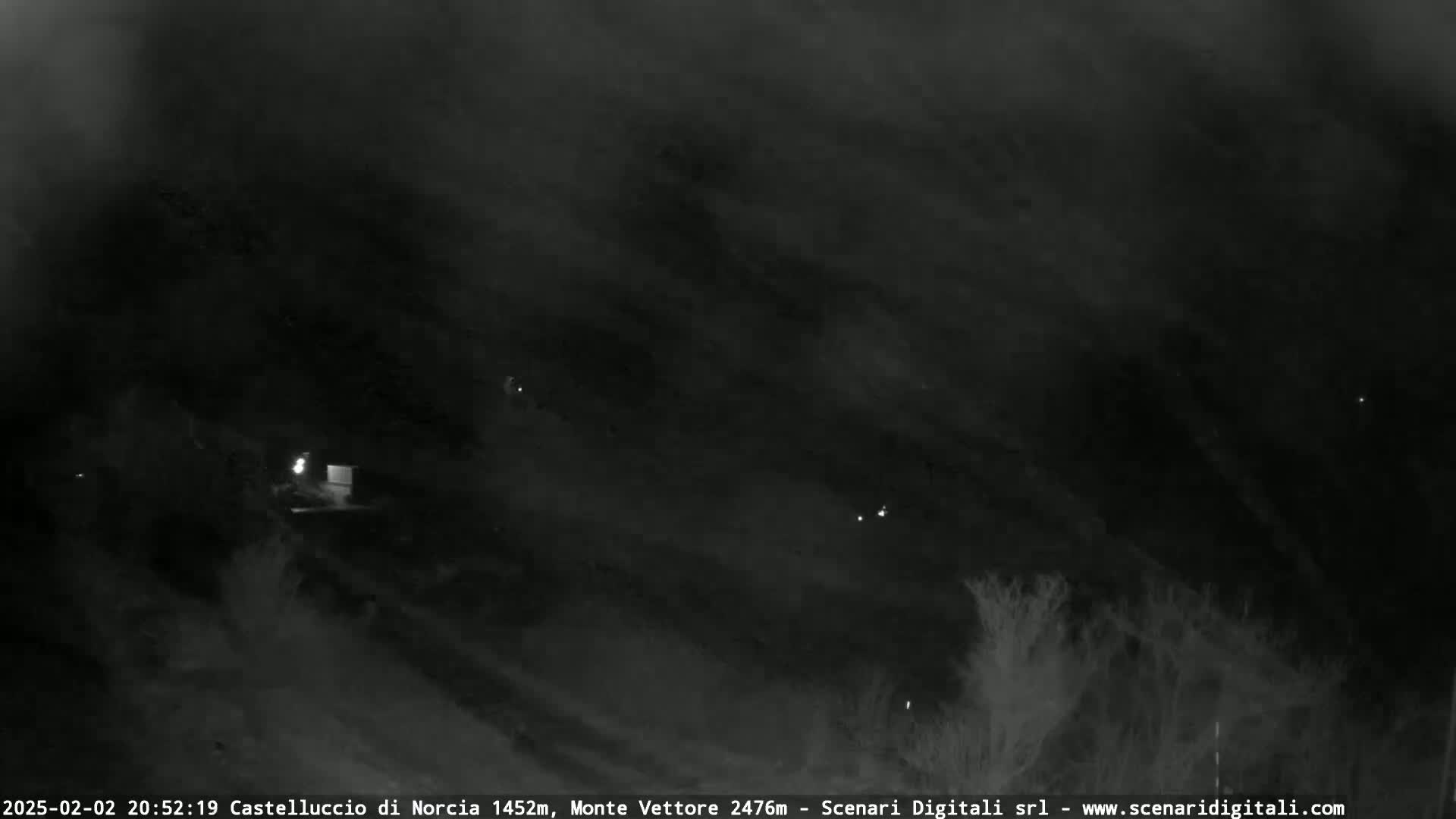 A nighttime, low-light image shows a dark, possibly cloudy or hazy sky over a landscape with sparse vegetation and faint lights.