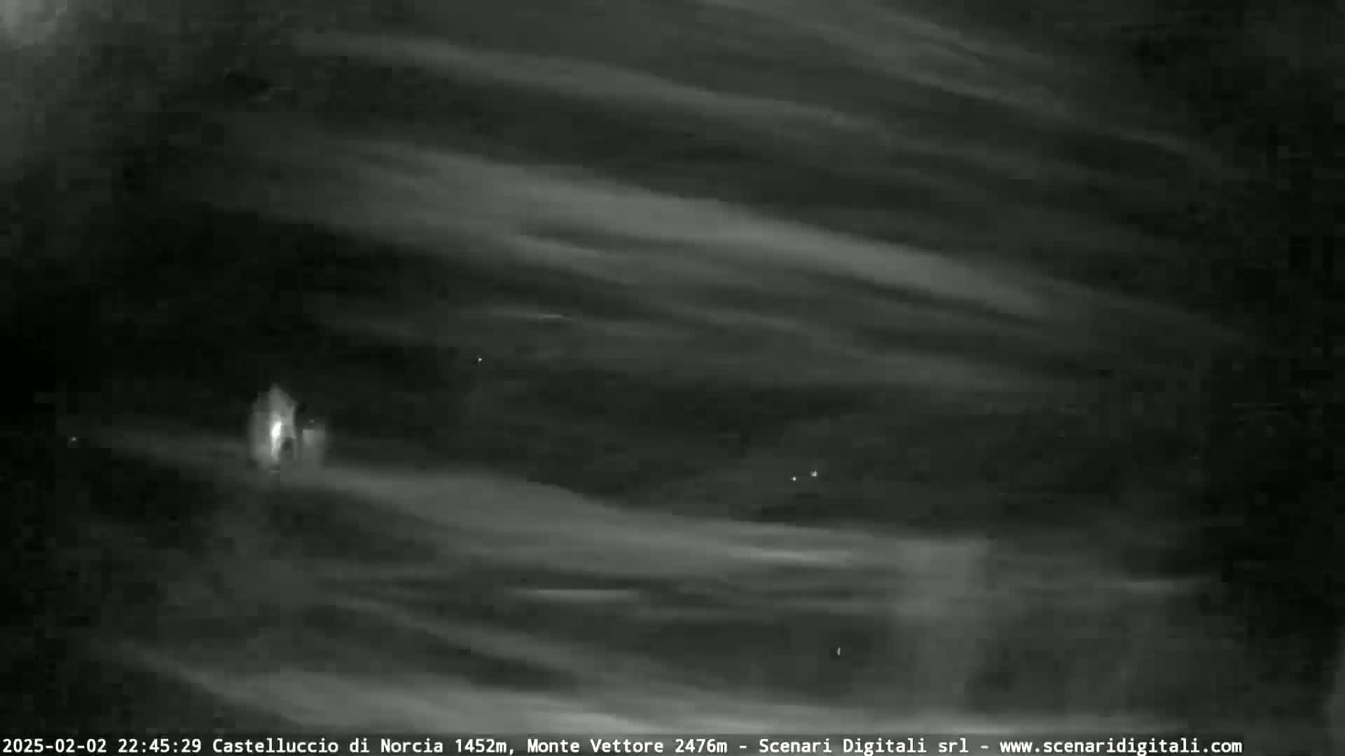 A nighttime long exposure shows thin, horizontal clouds streaking across a dark sky, with a single light source visible in the lower-center.